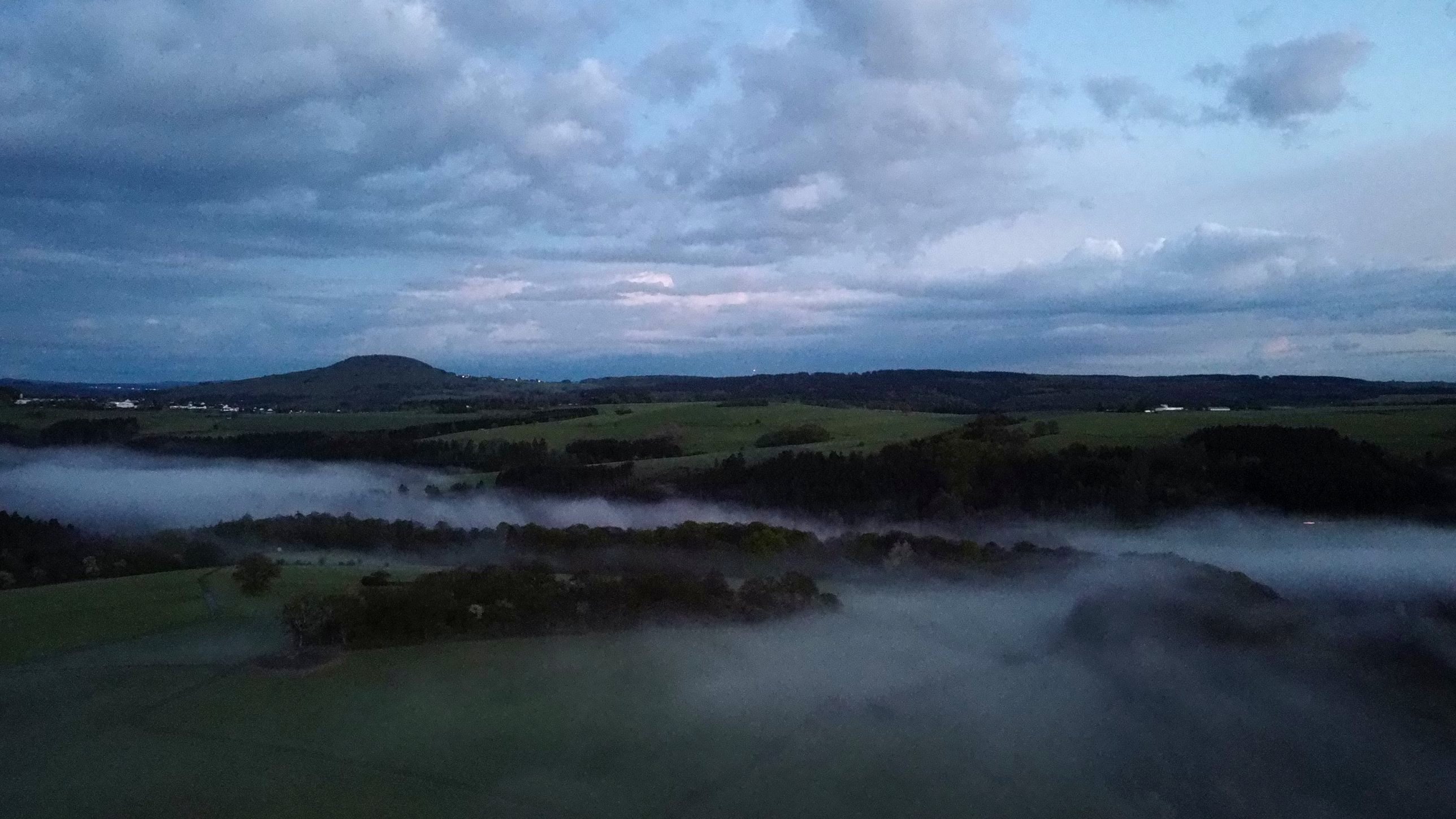 Nebelbänke über Wiesen und Wäldern bei Hümmel-Marthel in der Eifel, im Hintergrund ein Bergkegel, Drohnenaufnahme